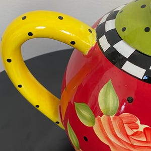 以下が含まれることがあります： 黒いドットが付いた黄色のハンドルが付いた、カラフルなセラミックティーポット。赤いボディには花の模様と緑の葉が描かれています。蓋は緑色で黒いドットがあり、白と黒のチェック柄です。