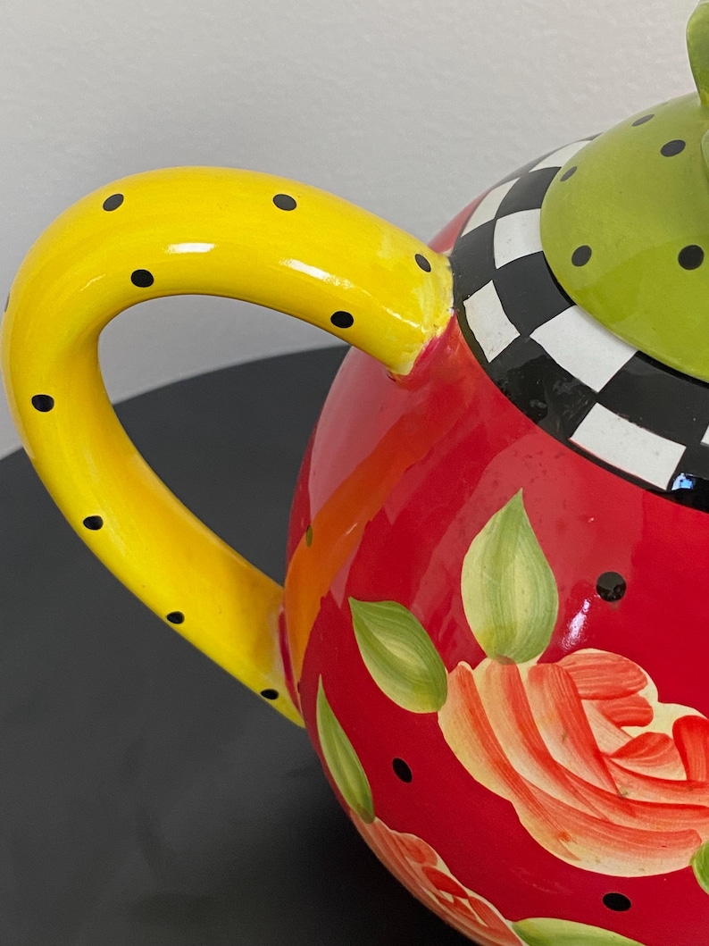 以下が含まれることがあります： 黒いドットが付いた黄色のハンドルが付いた、カラフルなセラミックティーポット。赤いボディには花の模様と緑の葉が描かれています。蓋は緑色で黒いドットがあり、白と黒のチェック柄です。