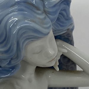 May include: A porcelain figurine of a woman with flowing blue hair, her face serene. The figure is in a relaxed pose, with one arm gently resting. The sculpture has a smooth, glossy finish, and the details are finely crafted.