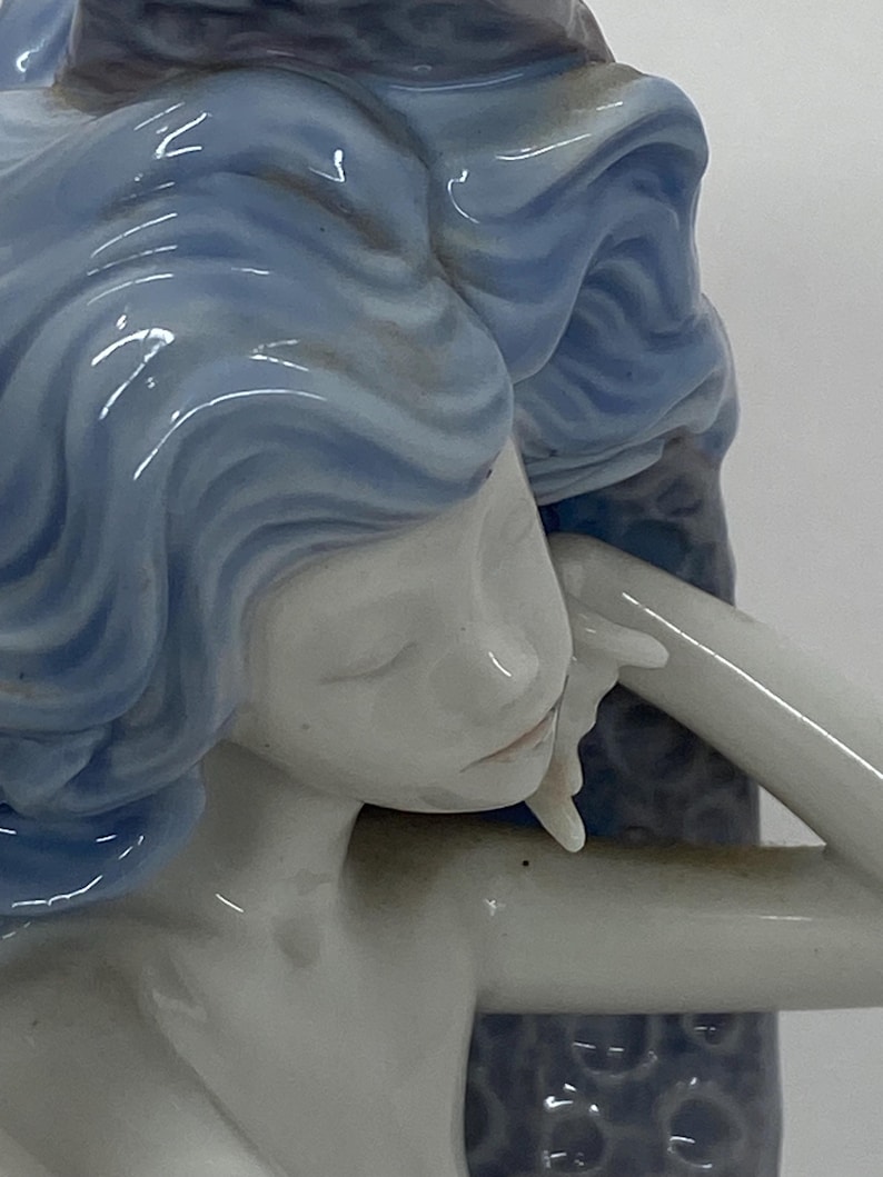 May include: A porcelain figurine of a woman with flowing blue hair, her face serene. The figure is in a relaxed pose, with one arm gently resting. The sculpture has a smooth, glossy finish, and the details are finely crafted.