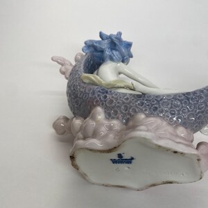 May include: A porcelain figurine of a mermaid reclining in a shell-shaped boat. The mermaid has blue hair and is resting on a lavender and pink base. The base has a textured pattern.