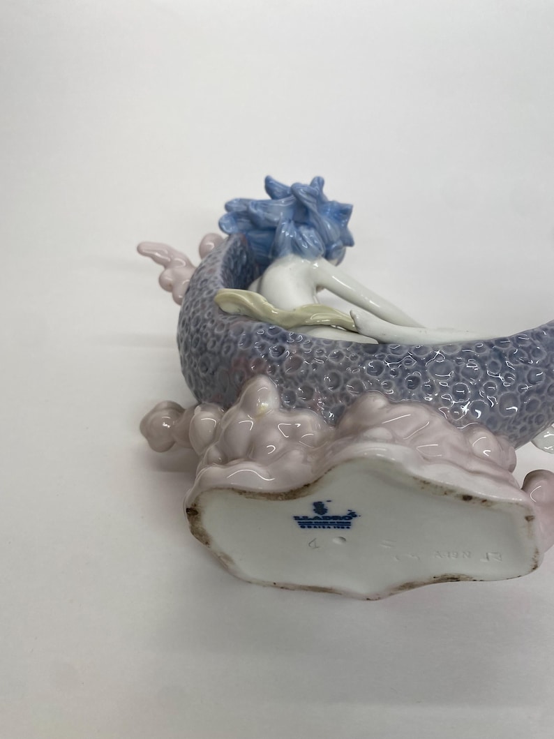 May include: A porcelain figurine of a mermaid reclining in a shell-shaped boat. The mermaid has blue hair and is resting on a lavender and pink base. The base has a textured pattern.