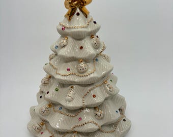 May include: A white ceramic Christmas tree figurine featuring a glossy finish, topped with a metallic gold bow. The tree is decorated with small, raised ornaments, gold beaded garland accents, and embedded colorful rhinestones throughout.