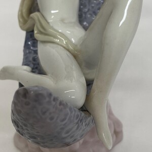 May include: A porcelain figurine of a seated figure with pale skin. The figure is draped with a light-colored cloth and seated on a textured, gray surface. The figurine has a delicate, detailed design and a glossy finish.
