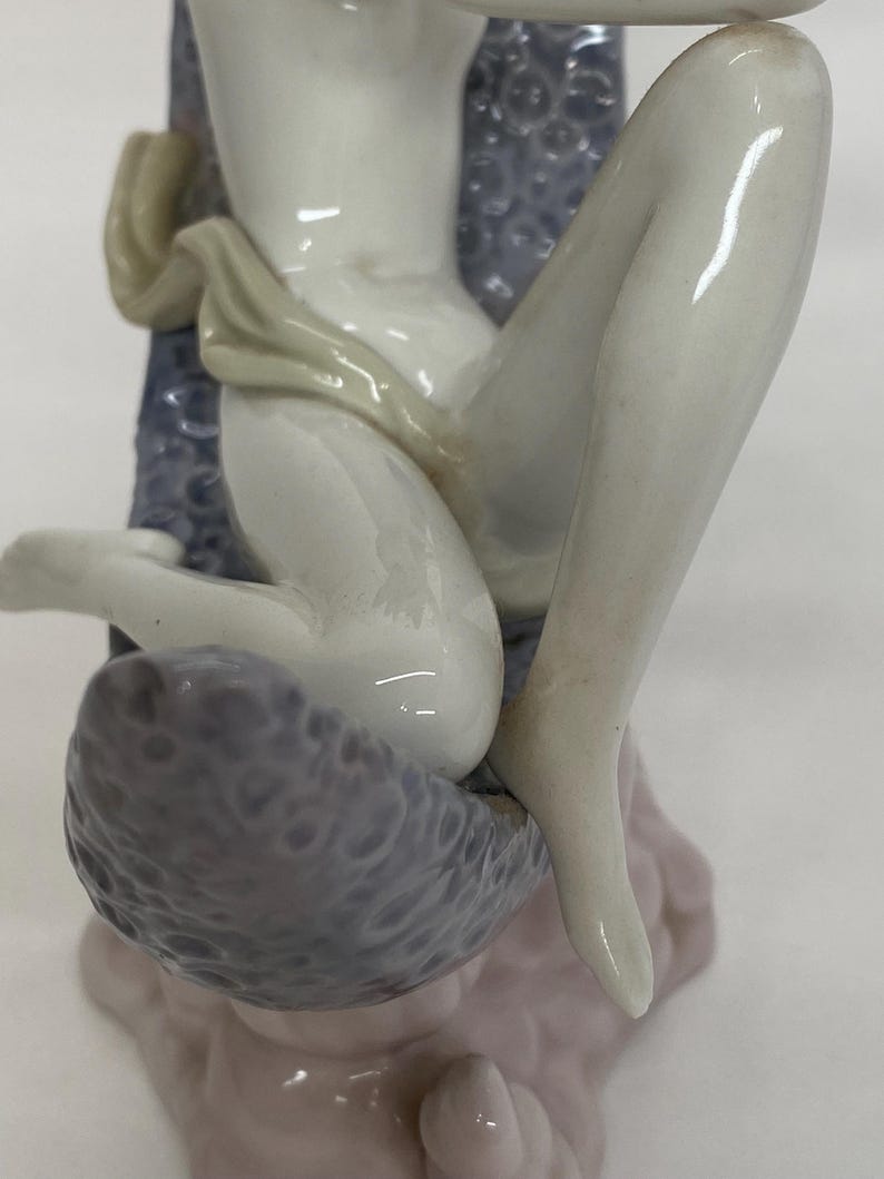 May include: A porcelain figurine of a seated figure with pale skin. The figure is draped with a light-colored cloth and seated on a textured, gray surface. The figurine has a delicate, detailed design and a glossy finish.