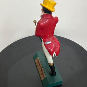 以下が含まれることがあります： 赤いコート、黄色のシルクハット、黒いブーツを履いた男性の置物。ステッキを持っています。置物は「Johnnie Walker」と書かれた緑色の台座の上に立っています。背景は白い壁と黒い表面です。