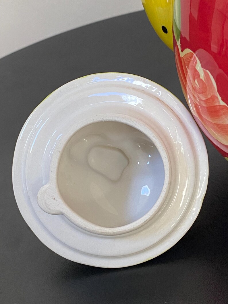 以下が含まれることがあります： 白いセラミックティーポットの蓋、取っ手付き。 蓋は丸く、縁が盛り上がっており、中央に開口部があります。 ティーポット本体は赤色で、花柄と黄色のアクセントが施されています。