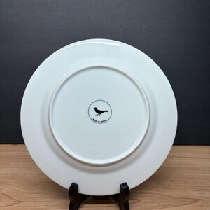Peut inclure: Une assiette en c&eacute;ramique blanche avec un logo d'oiseau noir et le texte "MADE IN JAPAN". L'assiette est pr&eacute;sent&eacute;e sur un support noir sur fond sombre. L'assiette a un design circulaire.