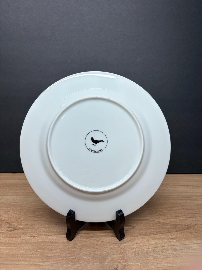 Peut inclure: Une assiette en c&eacute;ramique blanche avec un logo d'oiseau noir et le texte "MADE IN JAPAN". L'assiette est pr&eacute;sent&eacute;e sur un support noir sur fond sombre. L'assiette a un design circulaire.