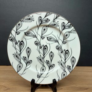 Peut inclure: Une assiette en c&eacute;ramique blanche avec un bord dor&eacute;, orn&eacute;e d'un motif floral noir et blanc. Le motif comprend des feuilles et des branches stylis&eacute;es. L'assiette est pr&eacute;sent&eacute;e sur un petit support noir, mettant en valeur son aspect d&eacute;coratif.