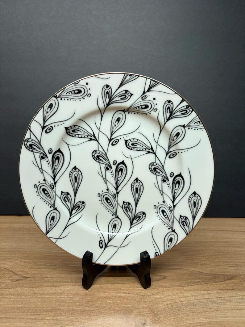 Peut inclure: Une assiette en c&eacute;ramique blanche avec un bord dor&eacute;, orn&eacute;e d'un motif floral noir et blanc. Le motif comprend des feuilles et des branches stylis&eacute;es. L'assiette est pr&eacute;sent&eacute;e sur un petit support noir, mettant en valeur son aspect d&eacute;coratif.