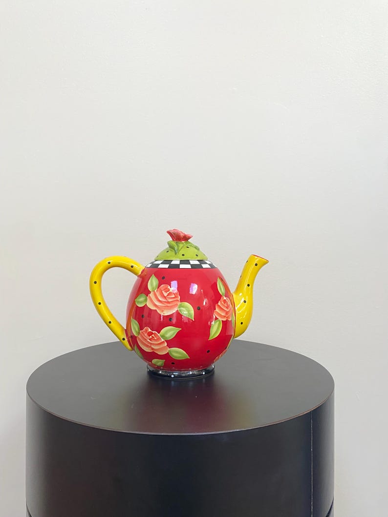 May include: A decorative teapot with a red body adorned with rose illustrations. The teapot features a yellow handle and spout with black polka dots, a green lid with a red rose, and a black and white checkered pattern on the rim.