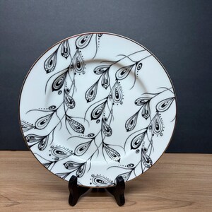 Peut inclure: Assiette en c&eacute;ramique blanche avec un motif floral noir et blanc. L'assiette a un bord dor&eacute; et est soutenue par un support noir. Le motif pr&eacute;sente des feuilles noires stylis&eacute;es et des formes de goutte d'eau.