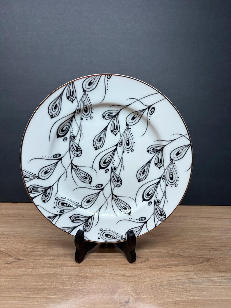 Peut inclure: Assiette en c&eacute;ramique blanche avec un motif floral noir et blanc. L'assiette a un bord dor&eacute; et est soutenue par un support noir. Le motif pr&eacute;sente des feuilles noires stylis&eacute;es et des formes de goutte d'eau.
