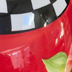 以下が含まれることがあります： 光沢のある赤い仕上げのセラミック容器のクローズアップ。容器は上部に黒と白のチェック柄、緑の葉のデザイン、白と赤のストライプ柄が特徴です。デザインは手描きです。