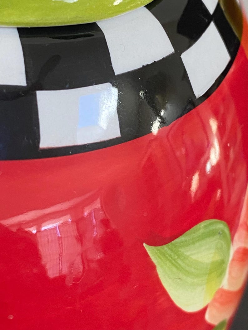 以下が含まれることがあります： 光沢のある赤い仕上げのセラミック容器のクローズアップ。容器は上部に黒と白のチェック柄、緑の葉のデザイン、白と赤のストライプ柄が特徴です。デザインは手描きです。