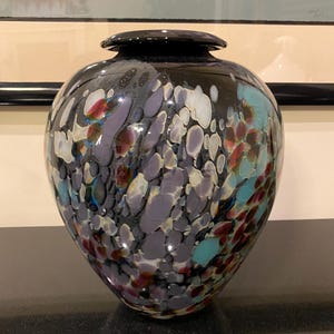 May include: A decorative glass vase with a black base and lid. The vase is adorned with a speckled pattern of purple, white, teal, and red. The vase has a rounded shape and sits on a black surface.