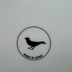 Peut inclure: Assiette en c&eacute;ramique blanche avec une silhouette d'oiseau noir &agrave; l'int&eacute;rieur d'un cercle noir. Les mots "MADE IN JAPAN" sont imprim&eacute;s en noir en dessous. L'assiette a une finition brillante.
