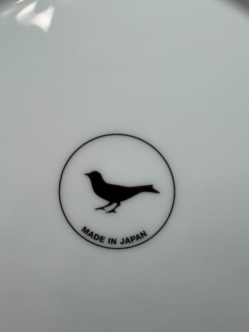 Peut inclure: Assiette en c&eacute;ramique blanche avec une silhouette d'oiseau noir &agrave; l'int&eacute;rieur d'un cercle noir. Les mots "MADE IN JAPAN" sont imprim&eacute;s en noir en dessous. L'assiette a une finition brillante.