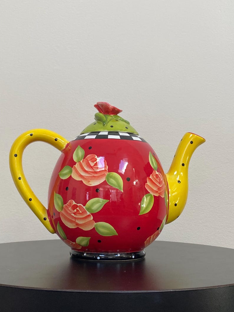 May include: A decorative red ceramic teapot with a yellow handle and spout. The teapot is adorned with orange roses, green leaves, and black polka dots. The lid is green with a red rose and a black and white checkered pattern.