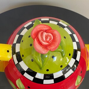 以下が含まれることがあります： 蓋に赤いバラが付いた装飾的なティーポット。 ティーポットは、花のアクセントが付いた赤いベース、黒い水玉模様の緑色の中心、白と黒の市松模様のボーダーを備えています。 黄色のハンドルがデザインを完成させています。