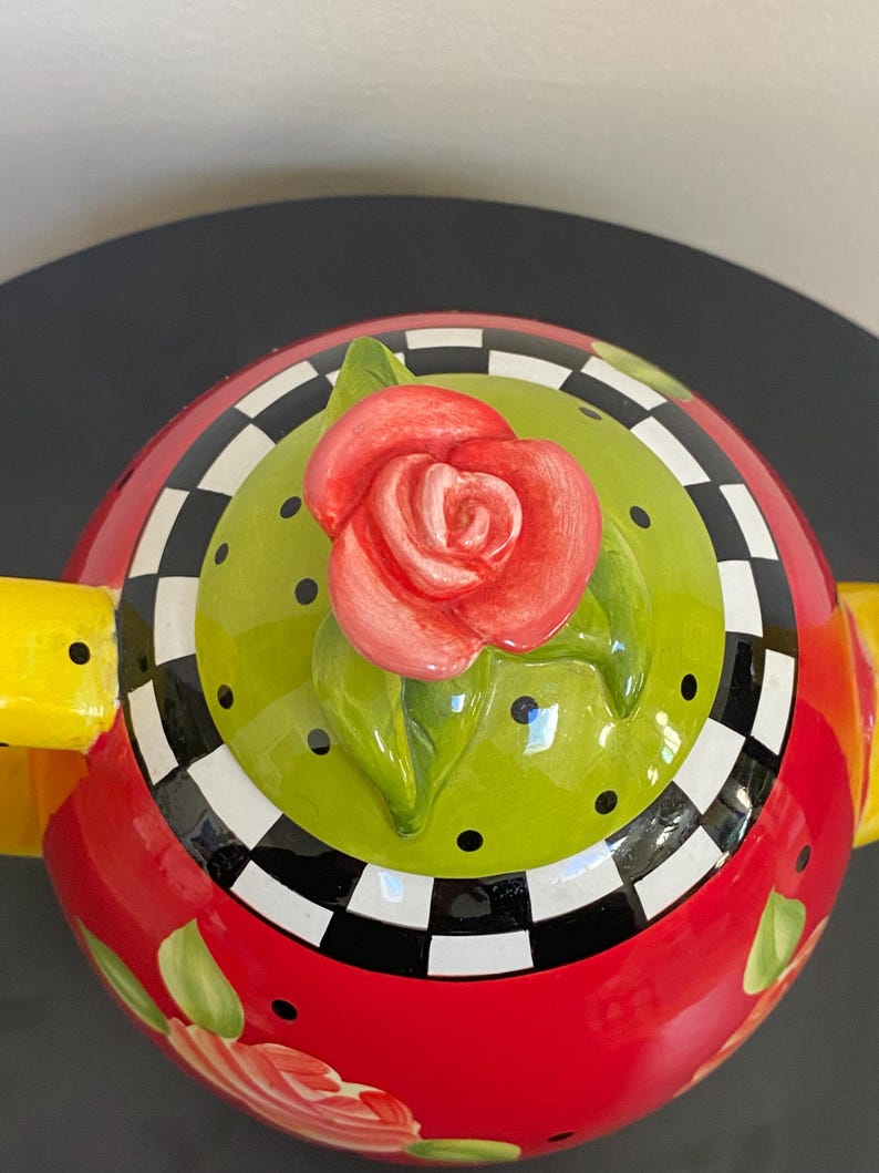 以下が含まれることがあります： 蓋に赤いバラが付いた装飾的なティーポット。 ティーポットは、花のアクセントが付いた赤いベース、黒い水玉模様の緑色の中心、白と黒の市松模様のボーダーを備えています。 黄色のハンドルがデザインを完成させています。