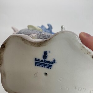 May include: A porcelain figurine with a blue and gray design. The base of the figurine is white with the blue "Lladro" logo and "Hand Made in Spain" text. The figurine has a detailed design.