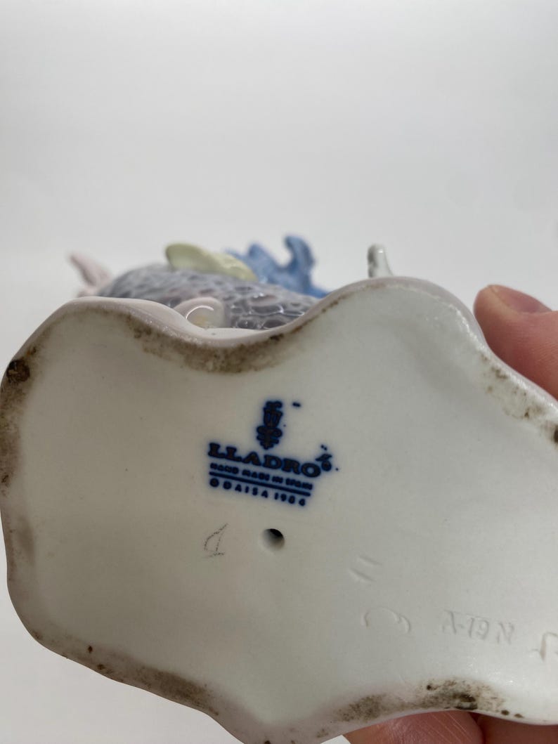 May include: A porcelain figurine with a blue and gray design. The base of the figurine is white with the blue "Lladro" logo and "Hand Made in Spain" text. The figurine has a detailed design.