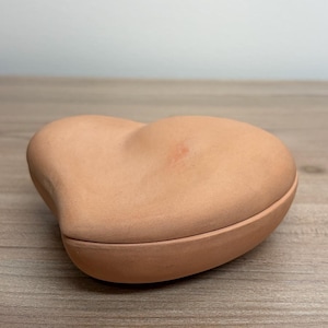 May include: A heart-shaped terracotta trinket box. The box is a warm, earthy tone, with a smooth, matte finish. The lid fits snugly onto the base, creating a seamless look. The box is sitting on a wooden surface.
