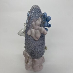 May include: A ceramic sculpture featuring a textured, gray-blue central form with a series of light pink and blue accents. The sculpture has a rounded shape with a textured surface and is set against a white background.