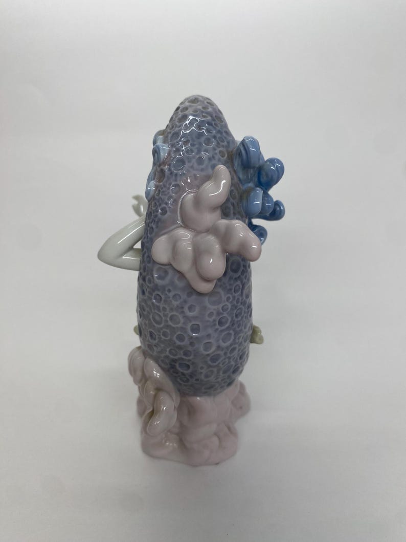 May include: A ceramic sculpture featuring a textured, gray-blue central form with a series of light pink and blue accents. The sculpture has a rounded shape with a textured surface and is set against a white background.