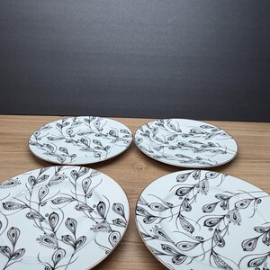 Peut inclure: Quatre assiettes ovales blanches avec un motif de feuilles noir et blanc et des bords dor&eacute;s. Les assiettes sont dispos&eacute;es sur une surface en bois, avec un fond gris fonc&eacute;.