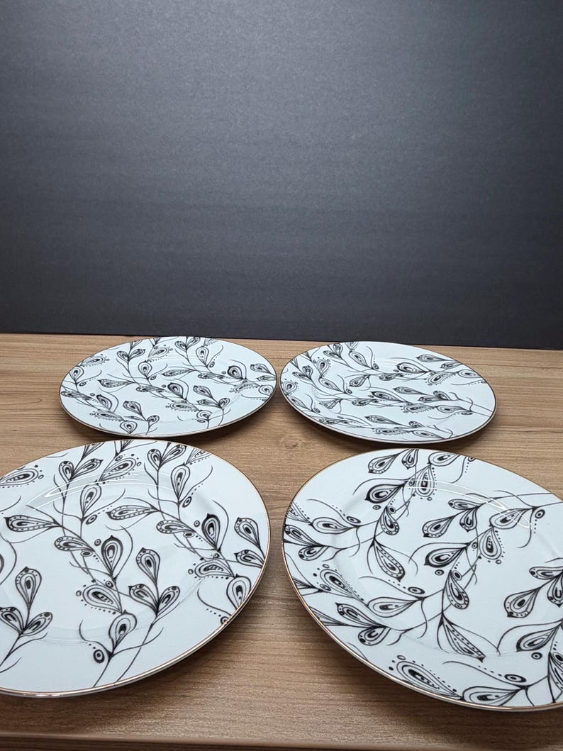 Peut inclure: Quatre assiettes ovales blanches avec un motif de feuilles noir et blanc et des bords dor&eacute;s. Les assiettes sont dispos&eacute;es sur une surface en bois, avec un fond gris fonc&eacute;.