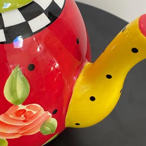 以下が含まれることがあります： カラフルな陶器のティーポット。赤いボディに花柄、黒い水玉模様の黄色の注ぎ口、白と黒の市松模様の緑色の蓋が特徴です。装飾的なキッチンアイテムです。