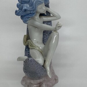 May include: A porcelain figurine of a seated figure with flowing blue hair. The figure is nude, with a light-colored cloth draped around the waist. The figure is seated on a textured, lavender-colored base, with a light pink base.