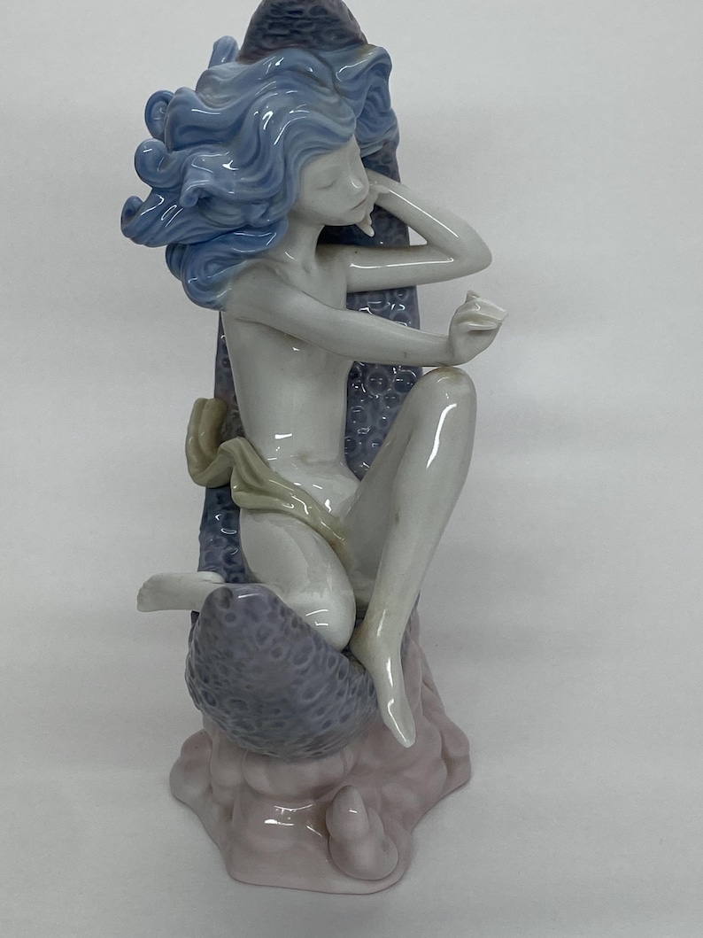 May include: A porcelain figurine of a seated figure with flowing blue hair. The figure is nude, with a light-colored cloth draped around the waist. The figure is seated on a textured, lavender-colored base, with a light pink base.