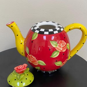 以下が含まれることがあります： 黄色い取っ手と注ぎ口が付いた赤いセラミックティーポット。黒い水玉模様と花の模様で装飾されています。蓋はライムグリーンで、赤いバラと黒い水玉模様が付いています。縁は白と黒のチェック柄です。