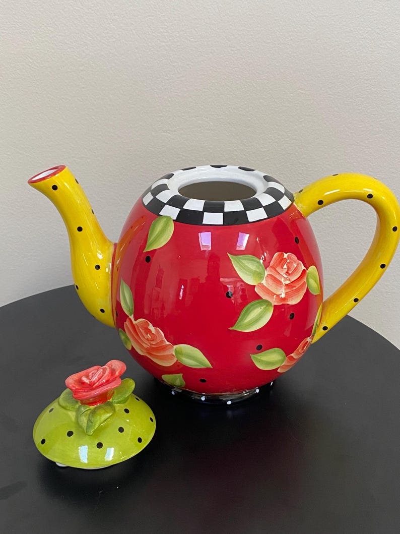 以下が含まれることがあります： 黄色い取っ手と注ぎ口が付いた赤いセラミックティーポット。黒い水玉模様と花の模様で装飾されています。蓋はライムグリーンで、赤いバラと黒い水玉模様が付いています。縁は白と黒のチェック柄です。