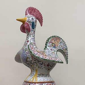 May include: A decorative ceramic rooster figurine. The rooster features a white base with colorful floral patterns, a red comb, and a yellow beak. The tail feathers are green with intricate designs. The figurine is on a pedestal base.