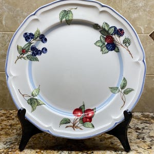 May include: White ceramic plate with a scalloped edge, adorned with a blue border and fruit illustrations. The plate features images of blueberries, raspberries, and cherries. The plate is displayed on a black stand.