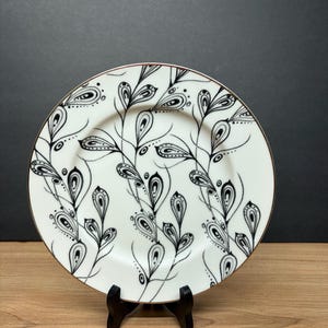Peut inclure: Assiette en c&eacute;ramique blanche avec un motif floral noir et blanc. L'assiette a un bord dor&eacute; et est pr&eacute;sent&eacute;e sur un pr&eacute;sentoir noir. Le motif comprend des formes de feuilles stylis&eacute;es et des lignes fines, cr&eacute;ant une esth&eacute;tique &eacute;l&eacute;gante.