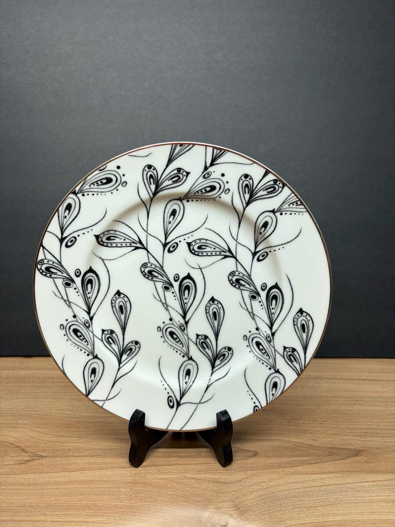 Peut inclure: Assiette en c&eacute;ramique blanche avec un motif floral noir et blanc. L'assiette a un bord dor&eacute; et est pr&eacute;sent&eacute;e sur un pr&eacute;sentoir noir. Le motif comprend des formes de feuilles stylis&eacute;es et des lignes fines, cr&eacute;ant une esth&eacute;tique &eacute;l&eacute;gante.