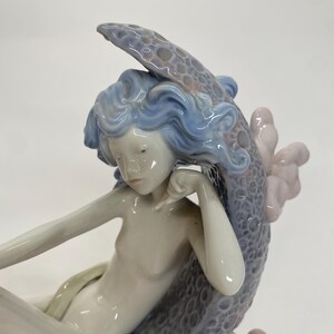 May include: A porcelain figurine of a mermaid with flowing blue hair and a serene expression. The mermaid is seated, with a snake coiled around her. The sculpture features a detailed, textured tail and is accented with soft pink and gray hues.