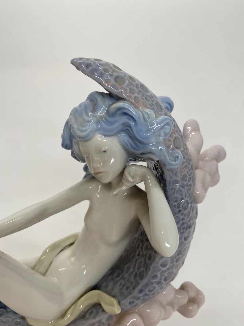 May include: A porcelain figurine of a mermaid with flowing blue hair and a serene expression. The mermaid is seated, with a snake coiled around her. The sculpture features a detailed, textured tail and is accented with soft pink and gray hues.