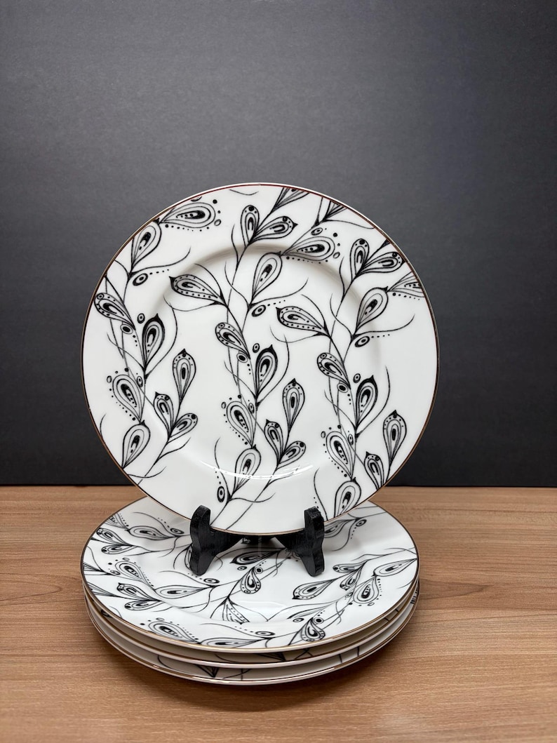 Peut inclure: Ensemble d'assiettes en c&eacute;ramique blanche avec un motif floral noir et blanc. Les assiettes ont un bord dor&eacute; et sont empil&eacute;es sur une surface en bois. Les assiettes sont pr&eacute;sent&eacute;es sur un support noir sur fond sombre.