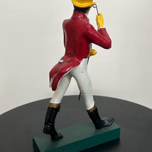 以下が含まれることがあります： 赤い燕尾服、黄色のシルクハット、黒いブーツを履いた人物の置物。 このフィギュアは鞭を持っており、黄色の手袋を着用しています。 フィギュアは緑色の台座の上に立っています。