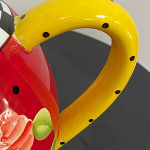 以下が含まれることがあります： 手描きのティーポットのクローズアップ。明るい黄色のハンドルには黒い水玉模様が施されています。ボディは鮮やかな赤色で、花の模様が描かれています。蓋の一部には、白と黒の市松模様があります。