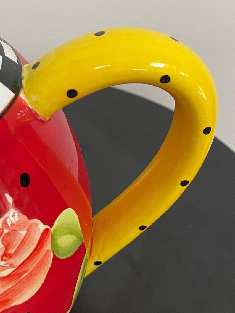 以下が含まれることがあります： 手描きのティーポットのクローズアップ。明るい黄色のハンドルには黒い水玉模様が施されています。ボディは鮮やかな赤色で、花の模様が描かれています。蓋の一部には、白と黒の市松模様があります。