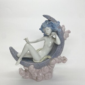 May include: A porcelain figurine of a reclining figure with blue hair, resting on a crescent moon. The moon is a textured gray-blue, supported by pink cloud-like formations. The figure is nude, with a serene expression.
