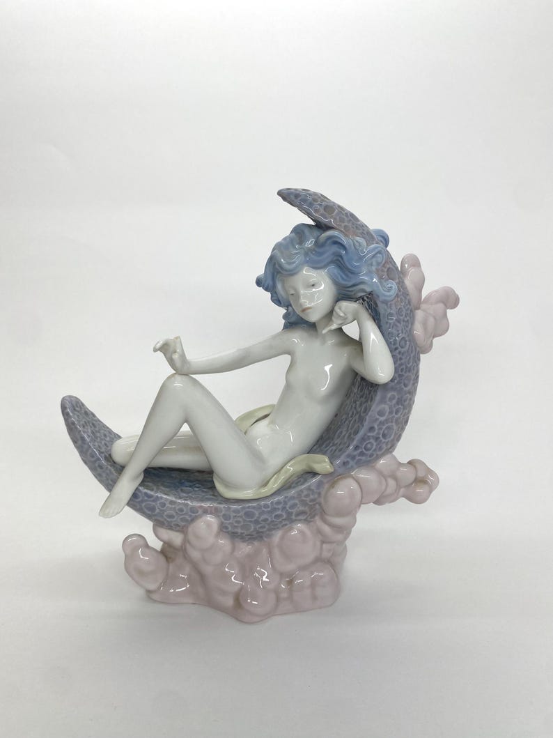 May include: A porcelain figurine of a reclining figure with blue hair, resting on a crescent moon. The moon is a textured gray-blue, supported by pink cloud-like formations. The figure is nude, with a serene expression.